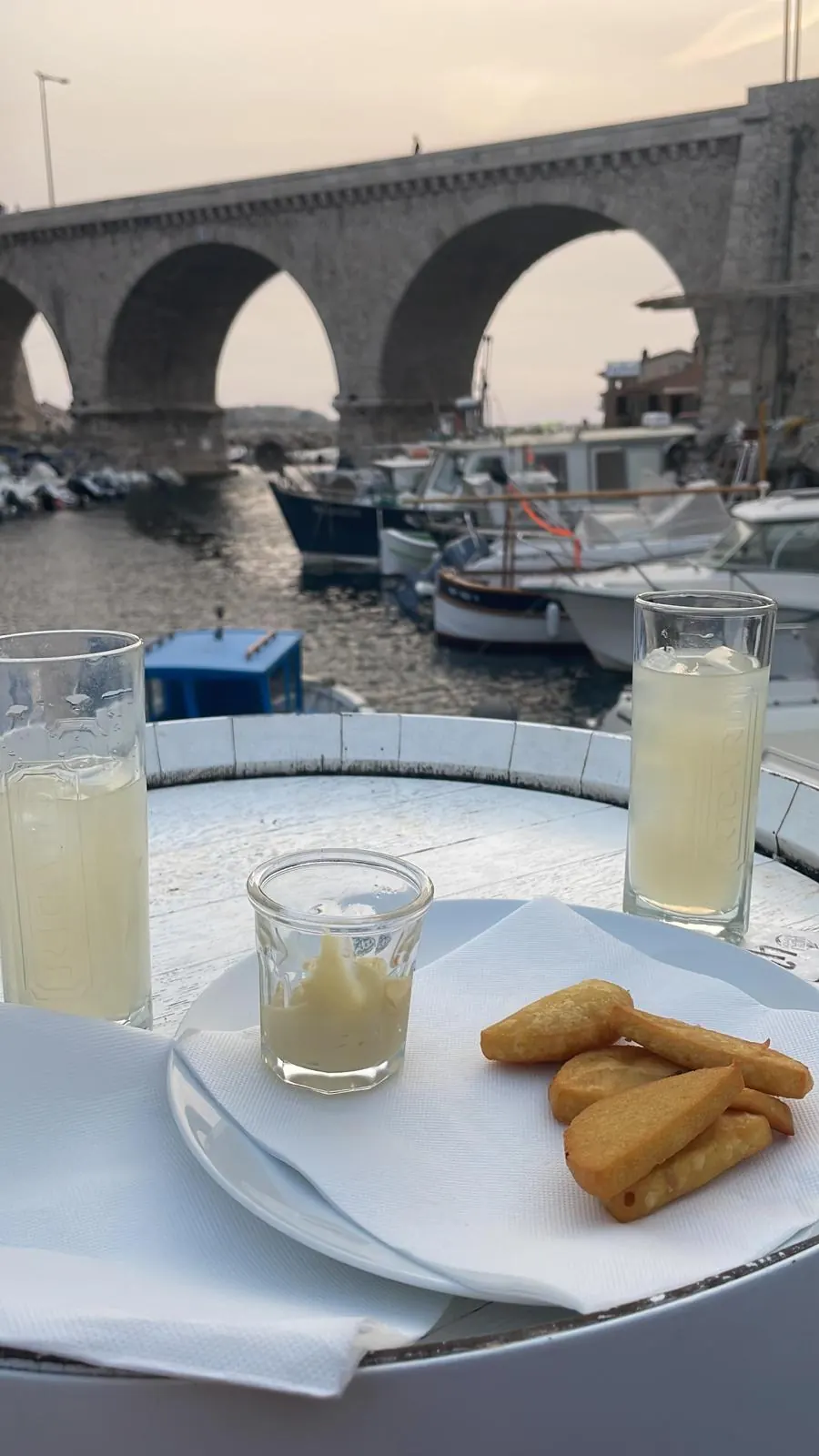 Pastis und Panisses auf einem Tisch im Vallon des Auffes, dahinter Boote und das Viadukt